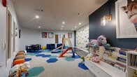 This thoughtfully designed children's playroom is a single-level space packed with engaging features that cater to young, active minds. The room is anchored by a striking full-wall rock climbing installation fitted with colorful handholds, offering a challenging and exciting physical activity for children of various skill levels. Complementing this is a freestanding wooden jungle gym complete with a slide, climbing rungs, and rope elements, all situated atop a vibrant, hexagonal-patterned carpet in bold shades of blue and teal that defines the active play zone.

The space transitions seamlessly into quieter activity areas, including a child-sized table and chair set ideal for arts, crafts, or games, flanked by a row of playful mushroom-shaped stools in orange and teal. A cozy seating nook with a plush navy sofa provides a comfortable retreat. A dedicated toy and book storage area features a white shelving unit stocked with stuffed animals, books, and play sets, set against a dramatic deep navy accent wall illuminated by warm pendant lighting.

Bright recessed lighting throughout keeps the entire room well-lit, while vibrant, animal-themed artwork — including bold prints of a zebra, elephant, and giraffe — adds a lively, safari-inspired aesthetic. The clean white walls and neutral carpet outside the play zones maintain a fresh, organized atmosphere throughout.