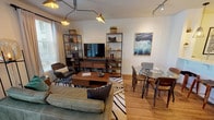 This thoughtfully designed single-floor residence blends mid-century modern style with contemporary comfort throughout its open, cohesive layout. The living area serves as the heart of the space, showcasing warm hardwood-style flooring, a striking multi-arm pendant light fixture, and flanking open metal-and-wood shelving units that frame a central media console. A plush sofa anchors the seating area over a bold geometric area rug, while natural light streams in through large windows dressed with linen curtains and blinds. A round glass dining table with wooden chairs sits adjacent, complemented by a framed ocean-wave artwork that adds a coastal touch. The kitchen features gray shaker-style cabinetry, a white quartz peninsula with an undermount sink, stainless steel appliances including a gas range, microwave, and French door refrigerator, and industrial-style pendant lighting overhead. The bedroom is a standout, featuring a rich teal accent wall adorned with a gallery of framed artwork, a ceiling fan, and floor-to-ceiling glass doors opening to a private outdoor space. The bathroom is clean and modern, with a blue-gray vanity, white quartz countertop, a large mirror, and light tile flooring, accented with playful framed art. Every detail reflects a curated, design-forward sensibility throughout.