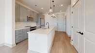 Located at 341 Great Circle Rd in Nashville, Tennessee, this single-floor unit offers a clean, contemporary design throughout. The open-concept living area is finished with light wood-look flooring, crisp white walls, and a ceiling fan, while a large window and glass-panel door flood the space with natural light and provide views of the surrounding cityscape. The kitchen is a standout feature, showcasing grey shaker-style cabinetry with gold hardware, a mosaic tile backsplash, stainless steel appliances including a spacious refrigerator, and a generous white quartz island with an undermount sink and stylish pendant lighting — making it both functional and visually striking. The bedroom is generously sized, carpeted for comfort, and benefits from dual windows that invite abundant natural light and ceiling fan. An additional room, also carpeted and sun-drenched, offers flexible use as a second bedroom or home office. The bathroom is elegantly appointed with a dual-sink vanity featuring grey cabinetry, gold hardware, illuminated mirrors, white quartz countertops, and light wood-look flooring, complemented by a full soaking tub. Throughout the unit, the consistent use of neutral tones and modern finishes creates a bright, cohesive, and inviting atmosphere.