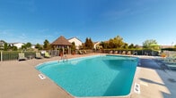 Located at Parkway Village in Grove City, Ohio, this outdoor amenity area serves as a central recreational hub for the community. The centerpiece is a generously sized swimming pool with crystal-clear blue water, framed by a wide concrete deck that provides ample space for lounging and socializing. The pool is designated as a no-diving pool and is accessed via built-in entry steps, making it accessible and family-friendly.

Surrounding the pool, a collection of green sling-style lounge chairs and seating arrangements invite residents to relax in the open air. A charming gazebo with a distinctive hip roof anchors one corner of the pool deck, offering a shaded retreat and a gathering spot for residents. Potted plants and greenery add decorative touches throughout the space, enhancing the welcoming atmosphere.

The perimeter of the pool area is enclosed by a metal fence, ensuring safety while maintaining an open feel with views of the surrounding landscaped grounds and mature trees. A children's playground is visible just beyond the fenced area, making this a well-rounded outdoor space suitable for all ages. The backdrop features the residential buildings of the Parkway Village community, reinforcing the neighborhood character of this amenity-rich outdoor environment.