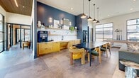 Located at 965 Duncan Perry Rd in Grand Prairie, Texas, this thoughtfully designed single-floor amenity space offers a well-rounded communal environment across a comfortable footprint. The interior is anchored by a welcoming lounge area featuring soaring ceilings with warm wood accents, a striking floor-to-ceiling white tile accent wall with a mounted flat-screen display, and an eclectic mix of stylish seating including a plush sectional, leather chairs, and sculptural side tables. Large black-framed floor-to-ceiling windows flood the space with natural light and provide open views of the surrounding community. Adjacent to the lounge, a game area houses a shuffleboard table set against expansive glazed walls, enhancing the bright, airy atmosphere. The café-style kitchenette is outfitted with warm wood cabinetry, a white marble-look backsplash, open shelving for décor and essentials, a coffee station, and a dark communal dining table with pendant lighting overhead. A private work nook provides a quiet retreat, featuring a sleek stone-top built-in desk, a cushioned swivel chair, and patterned carpeting underfoot. Polished large-format tile flooring runs throughout, tying together the modern, cohesive aesthetic of this versatile community hub.