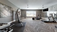This fitness center is thoughtfully designed to accommodate a wide range of workout needs within a single, open-plan space. The interior is finished with neutral gray walls, crisp white baseboards, and a distinctive herringbone-patterned carpet flooring that lends a polished, modern aesthetic throughout.

The cardio zone is well-stocked with treadmills, elliptical trainers, and a stair climber, complemented by a wall-mounted flat-screen television for entertainment during workouts. Full-length mirrors line one wall, visually expanding the space while allowing users to monitor their form. Ceiling fans provide additional air circulation throughout the main exercise area.

The strength training section includes a multi-station cable machine, an adjustable workout bench, and a comprehensive rack of dumbbells in varying weights, offering versatile options for resistance training. A rubber floor mat beneath the free weights adds safety and practicality.

A dedicated cycling area houses a spin bike and two upright stationary bikes, situated adjacent to a framed interior window that overlooks a separate children's play room — a convenient feature for families. Natural light filters in through multiple windows, offering views of the surrounding outdoor landscape and contributing to the bright, energizing atmosphere of the space.