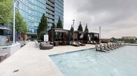This amenity space offers a compelling combination of outdoor recreation and indoor fitness facilities. The outdoor pool area is a standout feature, presenting a generously sized swimming pool with a smooth, light-toned deck. Along the poolside, a row of contemporary lounge chairs provides ample space for sunbathing and relaxation. Several sleek pergola-style cabanas line the pool's edge, each outfitted with slatted wood panels, pendant lighting, and plush wicker seating arrangements, offering shaded retreats. Mature cypress trees and curated landscaping add a refined, resort-like atmosphere, while the adjacent high-rise tower provides a dramatic urban backdrop.

The indoor fitness center is thoughtfully designed with a modern aesthetic, featuring patterned carpet flooring, recessed lighting, and floor-to-ceiling windows that invite natural light and outdoor views. The facility is comprehensively equipped with a full range of strength-training machines, free weights, benches, and cardio equipment including treadmills and ellipticals. A built-in millwork console with a white marble-style countertop, open shelving, and large-screen displays adds a polished, functional element to the space, bridging the entry corridor with the main workout floor.