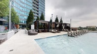 This amenity space offers a compelling combination of outdoor recreation and indoor fitness facilities. The outdoor pool area is a standout feature, presenting a generously sized swimming pool with a smooth, light-toned deck. Along the poolside, a row of contemporary lounge chairs provides ample space for sunbathing and relaxation. Several sleek pergola-style cabanas line the pool's edge, each outfitted with slatted wood panels, pendant lighting, and plush wicker seating arrangements, offering shaded retreats. Mature cypress trees and curated landscaping add a refined, resort-like atmosphere, while the adjacent high-rise tower provides a dramatic urban backdrop.

The indoor fitness center is thoughtfully designed with a modern aesthetic, featuring patterned carpet flooring, recessed lighting, and floor-to-ceiling windows that invite natural light and outdoor views. The facility is comprehensively equipped with a full range of strength-training machines, free weights, benches, and cardio equipment including treadmills and ellipticals. A built-in millwork console with a white marble-style countertop, open shelving, and large-screen displays adds a polished, functional element to the space, bridging the entry corridor with the main workout floor.