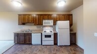 Located at 3755 Eighth Ave S in Saint Cloud, Minnesota, this single-floor unit offers a practical and comfortable layout designed for everyday living. The kitchen is well-equipped with rich, dark-stained wood cabinetry providing ample upper and lower storage, a double-basin sink, a dishwasher, a coil-top electric range, a built-in microwave, and a full-size refrigerator. The flooring in the kitchen features durable tile in a warm, earthy tone that complements the cabinetry. The kitchen opens toward the living area, which transitions to soft, neutral carpeting and is brightened by a window that allows natural light to filter in. A wall-mounted air conditioning unit provides climate control in this shared space, and baseboard heating runs along the perimeter. The bedroom is a comfortably sized, carpeted room featuring a ceiling fan with integrated lighting, a baseboard heater, and a window with an outdoor view. The bathroom includes a vanity with dark wood cabinetry, a white countertop, a lighted mirror, tile flooring consistent with the kitchen, and a shower/tub combination. Throughout the unit, neutral wall tones and consistent finishes create a cohesive, understated aesthetic well-suited for functional, day-to-day living.