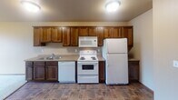 Located at 3755 Eighth Ave S in Saint Cloud, Minnesota, this single-floor unit offers a practical and comfortable layout designed for everyday living. The kitchen is well-equipped with rich, dark-stained wood cabinetry providing ample upper and lower storage, a double-basin sink, a dishwasher, a coil-top electric range, a built-in microwave, and a full-size refrigerator. The flooring in the kitchen features durable tile in a warm, earthy tone that complements the cabinetry. The kitchen opens toward the living area, which transitions to soft, neutral carpeting and is brightened by a window that allows natural light to filter in. A wall-mounted air conditioning unit provides climate control in this shared space, and baseboard heating runs along the perimeter. The bedroom is a comfortably sized, carpeted room featuring a ceiling fan with integrated lighting, a baseboard heater, and a window with an outdoor view. The bathroom includes a vanity with dark wood cabinetry, a white countertop, a lighted mirror, tile flooring consistent with the kitchen, and a shower/tub combination. Throughout the unit, neutral wall tones and consistent finishes create a cohesive, understated aesthetic well-suited for functional, day-to-day living.