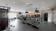 This fitness center is thoughtfully designed to accommodate a full range of workout needs within a spacious, single-level layout. The space is divided into distinct functional zones, beginning with a dedicated cardio area housing multiple treadmills, elliptical trainers, upright stationary bikes, and a rowing machine. Each cardio machine is equipped with an individual display console, and ceiling-mounted televisions are positioned throughout for entertainment during workouts. Ceiling fans provide consistent airflow across the room.

Large mirrors line the walls, visually expanding the space and enhancing the open feel. Bold orange accent walls add energy and visual contrast to the otherwise neutral grey-and-white color scheme. Durable rubber flooring covers the entire space, offering both safety and sound absorption.

A separate strength training area features a multi-functional cable machine, a tiered dumbbell rack with a broad range of free weights, and an adjustable workout bench. Stability balls and additional accessories are also available. Natural light filters in through multiple windows fitted with blinds, and recessed lighting supplements the brightness throughout. Crown molding along the ceiling adds a refined architectural touch to the overall design.