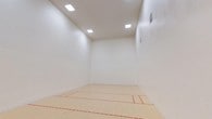 This racquetball court offers a fully enclosed, regulation-ready playing environment designed for optimal sport performance. The court is defined by smooth, white-painted walls on all four sides, providing consistent ball rebound essential to the game. The floor features a light-toned hardwood surface with clearly marked red boundary and service lines, adhering to standard racquetball court specifications.

The space rises to a generous ceiling height, giving players ample vertical room for play. Overhead, recessed skylight-style fluorescent panels provide bright, even illumination throughout the court, minimizing shadows and ensuring excellent visibility. Ventilation is maintained through wall-mounted HVAC vents positioned at multiple points around the court.

A single entry door with a small window is located on the back wall, providing access while maintaining the enclosed integrity of the playing environment. The overall ambiance is clean, functional, and purpose-built, with an uncluttered design that keeps the focus entirely on the playing surface. This court is well-suited for both casual play and competitive practice sessions.