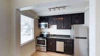 This single-floor residence in Gallery, US, offers a clean, minimalist aesthetic characterized by crisp white walls, white trim, and light hardwood flooring that flows consistently throughout the space. The open layout of the main living area creates a sense of spaciousness, with a structural beam subtly defining zones while maintaining visual continuity between rooms. Multiple doorways connect the living area to adjacent rooms, and a window at the far end of the hallway draws natural light deep into the interior.

The kitchen is a standout feature, designed with dark espresso-toned cabinetry that extends from floor to ceiling, providing generous storage capacity. Granite countertops in a complementary dark tone add texture and contrast, while a full suite of stainless steel appliances—including a range, over-the-range microwave, dishwasher, and refrigerator—offers modern functionality. Track lighting overhead illuminates the workspace effectively, and a large window beside the cabinetry floods the kitchen with abundant natural light. The light hardwood flooring continues here, tying the kitchen cohesively to the rest of the home.