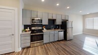 This single-floor unit showcases a contemporary open-concept design that seamlessly connects the kitchen and living areas. The kitchen is well-appointed with gray shaker-style cabinetry, granite-look countertops, a dark subway tile backsplash, and a full suite of stainless steel appliances including a range, microwave, dishwasher, and refrigerator. Wood-style plank flooring runs throughout the main living areas, adding warmth and continuity to the space.

The living area receives generous natural light through a large window and a sliding glass door that opens to a private balcony, offering an indoor-outdoor connection. Recessed lighting is featured throughout, providing consistent illumination across the open layout.

The bedroom area transitions to plush carpeting, creating a softer, more comfortable feel. Neutral wall tones throughout the unit contribute to a clean, versatile aesthetic.

The bathroom mirrors the unit's modern finishes, featuring gray cabinetry with a granite-look vanity top, a large mirror, wood-style flooring, and a shower-tub combination. The entry area provides a practical transition space with direct access from the front door into the main living zone.