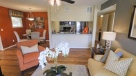 This thoughtfully designed single-floor residence offers an open-concept layout that seamlessly connects the living, dining, and kitchen areas into one cohesive and functional space. Rich hardwood flooring flows throughout, anchoring the warm, contemporary aesthetic.

The kitchen is positioned at the heart of the home, featuring stainless steel appliances — including a refrigerator, microwave, and range — complemented by granite countertops and dark cabinetry. A half-wall breakfast bar separates the kitchen from the living area, maintaining an open and airy feel while providing a natural division of space.

The living area is generously proportioned, accommodating comfortable seating arrangements beneath a ceiling fan with light fixture. Natural light filters in to brighten the space, creating a welcoming atmosphere. A plush area rug and tasteful furnishings complete the inviting feel of the room.

The dining area is distinguished by a bold terracotta accent wall and pendant lighting overhead, lending a warm, intimate character to mealtimes. Together, the connected spaces create a harmonious flow ideal for both everyday living and entertaining. The overall ambiance is modern yet warm, with rich tones and carefully considered details throughout.