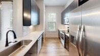 Located at 207 Gresham Ln in Mooresville, NC, this single-floor residence showcases a clean, modern design with a consistent palette of crisp white walls, warm wood-look flooring, and matte black hardware accents carried throughout. The galley-style kitchen is well-appointed with dark cabinetry, white quartz countertops, a hexagonal tile backsplash, and a full suite of stainless steel appliances including a refrigerator, range, microwave, and dishwasher. A window at the end of the kitchen allows natural light and outdoor views of the surrounding greenery. Adjacent to the kitchen, a dining or transition area features pendant lighting, a door opening to an exterior balcony, and additional windows that further brighten the space. The open living area offers recessed lighting and a spacious, versatile layout with clear sightlines to other areas of the home. Bedrooms are bright and airy, each featuring wood-look flooring, windows with outdoor tree views, and two-panel interior doors with black hardware. The primary bathroom includes a large framed mirror, white quartz vanity, dark cabinetry, a glass-enclosed walk-in shower with tile surround, and wood-look flooring. A secondary bathroom mirrors this refined aesthetic with a long vanity, open shelving, and coordinating fixtures throughout.