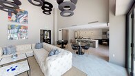 This contemporary communal space is designed with a seamless open-plan layout that encourages social interaction and relaxed gatherings. The centerpiece of the lounge area is a floor-to-ceiling built-in media wall, finished in warm grey wood tones and fitted with open shelving, drawers, and a mounted flat-screen TV. Dramatically high ceilings amplify the sense of openness, accentuated by a striking cluster of large circular pendant light fixtures that serve as a bold architectural focal point overhead. The living area is furnished with a generously sized tufted sectional sofa and leather accent chairs arranged around a marble-topped coffee table, all anchored by a woven area rug over light herringbone-patterned tile flooring. An open kitchen features a large island with a crisp white quartz countertop, dark cabinetry with stainless steel appliances, and elegant hanging pendant lights. Adjacent to the kitchen, a cozy dining nook offers banquette seating in deep blue velvet alongside a round black glass table, illuminated by a sculptural globe pendant and framed by large windows that invite abundant natural light and views of the outdoors. Vibrant artwork adorns the walls, adding color and personality to the overall refined, modern aesthetic.