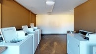 This single-floor shared laundry facility is designed to serve residents with both practicality and comfort in mind. The space is divided into two distinct functional zones, each contributing to a well-rounded communal experience.

The laundry area is equipped with multiple top-loading washing machines and dryers arranged along the walls, providing ample capacity for simultaneous use. Polished concrete flooring throughout the space lends an easy-to-maintain, durable surface that ties both zones together seamlessly. A wall-mounted air conditioning unit and baseboard heating ensure year-round climate comfort. Large horizontal windows admit generous natural light, offering views of the surrounding building exterior and greenery.

Adjacent to the laundry equipment is a dedicated seating lounge area, furnished with a leather armchair and a small side table, set atop a warm red area rug. Decorative metallic disc wall art adds a thoughtful aesthetic touch to the otherwise functional space. The warm tan and cream-toned walls contribute to an inviting, relaxed atmosphere.

Textured ceilings with flush-mounted lighting fixtures illuminate both zones evenly. The open layout and L-shaped floor plan allow for easy movement between the seating and laundry areas, making this a convenient and comfortable shared amenity space.