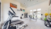 This fitness center is a thoughtfully designed exercise space housed within a single open room that makes an immediate impression with its soaring ceilings and an abundance of natural light. Large windows along one wall frame outdoor views, while clerestory windows near the roofline further brighten the crisp, white interior. The light-toned tile flooring complements the clean, modern aesthetic throughout.

The space is comprehensively outfitted to support a wide range of workout routines. Cardio equipment includes Spirit-brand treadmills and elliptical trainers, as well as a stationary cycling bike with a monitor. Strength training is well represented with a multi-station cable machine, a weight bench with a cable attachment system, a full dumbbell rack, and an assortment of stability and medicine balls. A large wall mirror on one side of the room enhances the sense of space and serves as a functional training aid.

Recessed ceiling lighting and a ceiling fan provide comfortable illumination and airflow, ensuring a pleasant environment during workouts. Vibrant sports-themed artwork adorns the upper walls, adding an energetic, motivational character to the room. A door provides access to an adjacent outdoor area, offering added convenience and connectivity to the broader facility.
