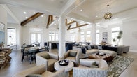 East Hall is a sophisticated single-story gathering space that blends transitional design with modern elegance. The interior is immediately distinguished by its soaring vaulted ceilings adorned with exposed wooden beam trusses, which contrast beautifully against the crisp white walls and structural columns to create a warm yet refined atmosphere.

The open-concept layout flows seamlessly between distinct functional zones. A generously sized lounge area anchors the main space, furnished with plush neutral-toned sofas, tufted leather ottomans, and curated accent chairs arranged to encourage both social interaction and relaxed conversation. Decorative table lamps and an orb-style pendant chandelier add layers of ambient lighting throughout.

Adjacent to the lounge, a dining and bar area features counter seating and darker, more intimate furnishings, offering a distinct contrast to the lighter lounge zone. Large windows lining the perimeter flood the interior with abundant natural light, enhancing the open and airy quality of the space.

Tasteful artwork, sculptural décor accents, and a cohesive neutral color palette with navy blue highlights complete the polished aesthetic, making East Hall a versatile and inviting communal destination.