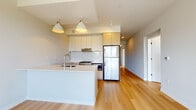 This thoughtfully designed single-floor residence showcases a clean, contemporary aesthetic with light hardwood flooring running throughout the open-plan living areas. The kitchen serves as a central focal point, featuring a spacious white island with an undermount sink, pendant lighting overhead, and a cohesive mix of white cabinetry accented by warm wood-toned upper cabinets. A white subway tile backsplash, gas range, built-in microwave, and stainless steel refrigerator complete the well-appointed culinary space. The open layout allows the kitchen to flow naturally into the living area, creating a bright and connected environment. A dedicated laundry closet houses a stacked full-size washer and dryer unit, adding everyday convenience. The bathrooms are finished in a crisp, minimalist style — one features a walk-in shower with floor-to-ceiling white surround and a chrome wall-mounted fixture, while the other offers a full soaking tub and shower combination. A sleek vanity with an undermount sink, white quartz countertop, and a large mirror completes the bathroom spaces. Throughout the residence, neutral tones and modern fixtures contribute to a cohesive, move-in-ready atmosphere.