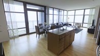 This single-floor space showcases a thoughtfully designed open-concept layout that integrates the kitchen, dining, and living areas into one cohesive environment. The kitchen features a substantial island topped with granite countertops and warm wood cabinetry with brushed metal hardware, providing ample storage and workspace. Light hardwood flooring extends throughout, lending warmth and continuity across the entire space.

The dining area accommodates a round glass-top table surrounded by upholstered chairs, positioned adjacent to floor-to-ceiling glass doors that open directly onto a balcony. The living area is furnished with a plush sofa set and accented by a patterned area rug, creating a comfortable and defined seating zone.

Expansive floor-to-ceiling windows wrap around the space, flooding the interior with abundant natural light and offering open outdoor views. The large glass panels and sliding doors provide a strong visual connection to the exterior balcony, enhancing the sense of openness. The overall ambiance is contemporary and airy, with clean lines, neutral tones, and modern finishes throughout contributing to a refined, functional living environment.