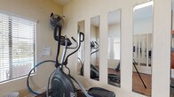 Located at 2937-2949 Unicornio St in Carlsbad, CA, this single-level fitness center is designed to provide residents with a dedicated space for cardiovascular exercise and general wellness. The room features warm wood-look flooring throughout, which adds a clean and polished aesthetic to the workout area.

The space is equipped with multiple pieces of cardio equipment, including an elliptical trainer, a stationary upright bike, and a recumbent bike, offering a variety of low-impact exercise options. A bold burnt-orange accent wall serves as a striking focal point, energizing the atmosphere and giving the space a gym-like character.

Mirror panels are strategically placed along multiple walls — including a set of tall vertical mirrors and a large framed mirror on the accent wall — which visually expand the room and allow users to monitor their form during workouts. Natural light filters in through windows fitted with horizontal blinds, keeping the space bright and well-ventilated.

A standing fan provides additional airflow, enhancing comfort during exercise. The entry door is conveniently located near a window, creating an airy feel. Overall, this fitness center is a practical and well-appointed amenity, thoughtfully designed to support an active lifestyle.