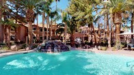 This inviting outdoor space presents a resort-style atmosphere centered around a sparkling turquoise swimming pool. The pool's most striking feature is a naturalistic rock waterfall formation, carefully constructed from stacked boulders that cascade water into the pool, creating a tranquil, resort-like ambiance. Towering palm trees and mature pines rise dramatically throughout the space, providing generous shade and a lush tropical aesthetic against the clear blue sky. The surrounding landscape is thoughtfully designed with additional rock accents, potted plants, and manicured greenery that enhances the tropical feel. A wrought iron fence borders portions of the area, adding a sense of definition and security without detracting from the open, airy environment. In the background, a multi-story Mediterranean-inspired building with warm earth-tone stucco walls and terracotta roofing ties the architectural aesthetic together cohesively. The overall ambiance is one of relaxation and leisure, offering a visually rich outdoor retreat that balances natural elements with well-maintained amenities. This space is ideal for those seeking a serene, resort-quality outdoor experience in a sun-drenched setting.