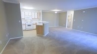 This single-floor residence offers a clean, open-concept layout that maximizes the sense of space and ease of movement throughout. The kitchen is well-appointed with white cabinetry, a white electric range, built-in microwave, and ample counter space along the perimeter. A peninsula-style island with a sink and warm wood-toned cabinetry serves as a natural divider between the kitchen and the expansive living area, providing additional prep and seating space while maintaining an open feel. The living area is finished with neutral-toned wall-to-wall carpeting that adds warmth and comfort underfoot. Soft, diffused overhead lighting illuminates the space evenly, and the light neutral wall color lends a calm, welcoming ambiance throughout. Crown molding adds a subtle architectural detail to the main living space. A glimpse into an adjoining room reveals a washer and dryer, indicating in-unit laundry convenience. The layout flows naturally from the kitchen through the living area and into additional rooms, making this a practical and comfortable single-level space well-suited for everyday living.