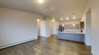 This single-floor space offers a clean, modern layout centered around an open-concept design that seamlessly connects the living area and kitchen. The vaulted ceilings add a sense of height and airiness to the main living space, which is finished with warm hardwood-style flooring that extends throughout. A baseboard heating unit runs along the living area wall, providing efficient climate control.

The kitchen is well-appointed with light wood cabinetry, a stainless steel refrigerator, and a kitchen island or peninsula finished in a contrasting blue-grey tone, topped with a light-colored countertop. Pendant lights hang above the kitchen area, adding focused task lighting and a stylish decorative touch. Recessed lighting throughout the ceiling further enhances the bright, welcoming ambiance.

Two paneled doors are visible from the main living space, leading to additional rooms or storage areas, suggesting a functional and organized floor plan. The neutral, warm grey wall palette complements the flooring and cabinetry, creating a cohesive and contemporary aesthetic. Overall, the space balances practicality with modern design, making it a comfortable and visually appealing single-level living environment.