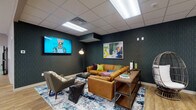 Located at 115 Carriage Hill Dr in Athens, Ohio, this single-floor community facility is thoughtfully designed to foster social interaction and comfort. The entry lobby sets a welcoming tone with light hardwood-style flooring, curated artwork, lush indoor greenery, and a mix of modern seating that invites residents and guests to gather.

The open dining and social area serves as the heart of the space, featuring light wood flooring, a statement pendant light fixture, round dining tables with an eclectic mix of teal and blue chairs, and a bold patterned accent wall. Multiple workstation desks line the perimeter, making this area functional as both a dining and co-working environment.

A dedicated lounge room offers a cozy retreat with dark teal textured walls, a large wall-mounted television, a warm cognac leather sectional sofa accented with colorful throw pillows, a wicker egg chair, and vibrant abstract artwork. The drop ceiling with recessed lighting gives the room a defined, intimate atmosphere.

The kitchen is clean and functional, outfitted with white cabinetry, dark countertops, stainless steel appliances including a full-size refrigerator, and a peninsula with seating. A connected computer workstation provides an added layer of utility. A well-lit corridor extends from the kitchen area, leading to additional rooms with keypad-secured entry doors, reinforcing the facility's organized and accessible layout throughout.
