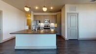 This two-floor residence offers a thoughtfully designed layout with a blend of modern finishes and functional living spaces. The open-concept main living area is anchored by a spacious kitchen island topped with a dark countertop and equipped with an integrated sink, illuminated by a trio of globe pendant lights. The kitchen itself features taupe cabinetry, a full suite of stainless steel appliances including a refrigerator, range, and microwave, complemented by a tile backsplash and under-cabinet lighting. Dark hardwood flooring flows throughout the main living areas, reflecting natural light that pours in through multiple large windows offering expansive waterfront views. A ceiling fan services the open living space, which also provides direct access to an outdoor balcony. The bathrooms are well-finished, with one featuring large-format tile flooring, a soaking tub with tiled surround, and an overhead shower, while another offers a walk-in glass-enclosed shower, a dark-cabinetted vanity with a backlit mirror, tile flooring, and access to a walk-in closet. The additional rooms are carpeted, bright, and airy, each featuring ceiling fans and windows — some with charming arched tops — that provide views of the surrounding community.