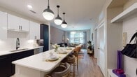 Located at 600 Willard St in Durham, NC, this single-floor residence offers a thoughtfully designed open-concept layout with contemporary finishes and a cohesive modern aesthetic throughout. The kitchen is a standout feature, showcasing a striking two-tone design with white upper cabinetry and dark lower cabinets, a white quartz waterfall island with bar seating, stainless steel appliances, and bold pendant lighting overhead. Light hardwood-style flooring extends seamlessly from the kitchen into the living and dining areas, creating a unified, airy feel. The living room is bathed in natural light from oversized windows and features warm, neutral tones accented by a rich teal sofa, an elegant chandelier, and a cozy dining nook positioned directly beside the window. The bedroom is a calm retreat with plush carpeting, a dramatic dark accent wall adorned with modern artwork, and a window with outdoor views. The bathroom is clean and refined, featuring a walk-in glass-enclosed shower with white subway tile, matte black hardware, and a white vanity with ample storage. Additional highlights include in-unit laundry and a functional entryway with built-in storage, making this residence as practical as it is stylish.
