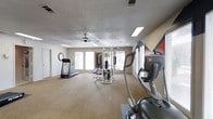 This single-floor fitness center offers a dedicated space for exercise and wellness. The room is generously sized, providing ample space for multiple users to work out comfortably without feeling crowded. Carpeted flooring spans the workout area, while a textured ceiling with a ceiling fan ensures air circulation throughout the space.

Natural light flows in through multiple large windows along the walls, creating a bright and energizing atmosphere. The room is further illuminated by recessed fluorescent lighting overhead.

The fitness center is outfitted with a well-rounded selection of equipment to support both cardiovascular and strength training workouts. Cardio options include a treadmill and an elliptical machine, while a multi-station cable and weight machine serves as the centerpiece for strength training exercises. An analog clock is mounted on the wall, allowing users to easily track their workout time. Motivational wall art in bold tones adds a dynamic visual element to the space.

Overall, the fitness center presents a clean, functional, and accessible environment suited for a range of fitness routines, making it a practical amenity for regular use.