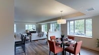 This single-floor residence offers an inviting open-concept design that effortlessly connects the dining and living areas into one cohesive, functional space. Rich hardwood flooring runs throughout, grounding the layout with warmth and a sense of continuity. A series of large windows lines the walls, flooding the interior with generous natural light and offering pleasant views of surrounding greenery.

The dining area is anchored by a round dark-topped table accompanied by bold terracotta-toned upholstered chairs, creating a striking focal point. Above, a contemporary drum pendant light adds an elegant finishing touch. A low half-wall partition subtly defines the transition between the dining and living zones without interrupting the open feel of the space.

The living area features a clean, modern aesthetic with a plush white sofa and coordinating accent pieces arranged on a patterned area rug. Tasteful decorative elements, including sculptural accents and soft lamp lighting, contribute to the refined yet comfortable ambiance. The neutral color palette of soft whites and warm grays throughout the space provides a versatile backdrop that feels both sophisticated and welcoming. Overall, this space combines practical design with an airy, light-filled atmosphere ideal for everyday living and entertaining.