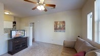This single-floor residence offers a practical and comfortable layout designed for everyday living. The living area is finished with soft wall-to-wall carpeting that adds warmth underfoot and a cozy ambiance throughout the space. A ceiling fan with an integrated light fixture is centrally mounted, providing both air circulation and ambient lighting to keep the room comfortable year-round. The neutral, light-colored walls create a clean and versatile backdrop, easily adaptable to a variety of décor styles. A decorative canvas print depicting an autumnal forest scene adds a touch of nature-inspired character to the room. The space is thoughtfully arranged to accommodate seating and entertainment furniture, with a dark wood media console positioned near the entryway wall. An open half-wall partition provides a subtle architectural division between the living area and an adjoining hallway, offering a sense of openness while maintaining a degree of spatial definition. The hallway beyond leads to additional rooms, suggesting a well-connected interior layout. Overall, the space presents a straightforward, move-in-ready environment with functional features and a calm, neutral aesthetic suited to a range of lifestyles.