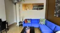 This single-floor space showcases a compelling blend of industrial and modern design elements. The living area is anchored by a striking royal blue L-shaped sectional sofa, dressed with an eclectic mix of decorative pillows in rich earth tones and botanical patterns, creating a vibrant focal point in the room. A sleek dark coffee table sits at the center, styled with glassware and a decorative tray, adding a touch of curated elegance.

One of the most distinctive architectural features is the large rectangular interior window cut into the upper portion of the wall, which reveals an adjacent space with exposed structural elements and warm ambient lighting, lending the overall layout an open, loft-like character. Exposed ductwork near the ceiling further reinforces the industrial aesthetic.

The flooring appears to be polished concrete, a hallmark of contemporary loft-style spaces, contributing to the cool, modern atmosphere. A side table with a sculptural branch arrangement adds an organic touch, softening the harder industrial elements. The overall ambiance is creative and modern, with layered textures and bold color choices defining the character of the space.