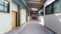This single-floor space showcases a thoughtfully designed interior corridor that merges industrial and historic architectural styles. The hallway stretches toward a distinctive arched brick passageway at its far end, lending a sense of historic character to the space. Overhead, exposed metal ductwork runs the length of the ceiling alongside skylights that draw in generous amounts of natural light, brightening the corridor throughout the day. Wall-mounted gooseneck light fixtures line both sides of the hallway, adding a utilitarian yet stylish complement to the industrial aesthetic. The flooring transitions between neutral carpet and stone tile, defining different zones along the corridor. The walls are finished in soft sage green with neutral trim, contributing to a calm, professional ambiance. Windows with dark frames are set into the right-hand wall, allowing additional light to filter through. Wooden doors with simple, clean hardware line the left side of the hallway, suggesting individual rooms or offices. The overall atmosphere balances historic architectural charm with practical, modern design elements, making it a functional and visually engaging space.