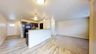 This single-floor space showcases a thoughtfully designed open-concept layout that seamlessly connects the kitchen and living areas. The kitchen is appointed with dark espresso-toned cabinetry, black appliances including a refrigerator, range, and overhead microwave, and a peninsula-style breakfast bar that serves as a natural divider between the kitchen and the main living space. Pendant lighting above the bar adds a warm, inviting ambiance, complemented by flush-mount ceiling fixtures throughout. The kitchen features smooth light-toned countertops that contrast elegantly with the dark cabinetry. The flooring transitions from wood-look planks in the kitchen and entryway to plush carpet in the living area, creating distinct zones within the open floor plan. Neutral gray walls and white trim provide a clean, versatile backdrop throughout the space. A hallway visible from the living area leads to additional rooms, with a paneled door offering privacy beyond. The overall atmosphere is modern yet cozy, with a practical layout well-suited for both everyday living and entertaining.