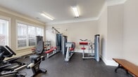 Located at 114 Avalon Park Rd in Mooresville, NC, this dedicated fitness center offers a well-equipped, single-floor workout facility designed to support a wide range of exercise routines. The space is finished with warm neutral walls, crown molding, and durable rubber flooring that provides both comfort and safety during workouts.

Natural light fills the room through multiple large windows fitted with horizontal blinds, offering views of the surrounding community and creating an open, inviting atmosphere. Overhead fluorescent lighting ensures the space remains well-lit throughout.

The cardio section is comprehensively stocked, featuring multiple treadmills equipped with individual display screens, an elliptical trainer, a stationary recumbent bike, and a rowing-style machine. A wall-mounted television is positioned for easy viewing during cardio sessions. The strength-training area includes a multi-station cable machine, a leg press and curl machine, a weight bench with press attachments, and a dumbbell rack offering a range of free weights.

Additional amenities include a wooden bench along the wall for use before or after workouts, a water dispenser, and a paper towel station for convenience and hygiene. Entry is provided through elegant French doors, adding a refined architectural touch to the functional space. The layout is thoughtfully organized to allow comfortable movement between equipment throughout the facility.