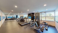 This single-floor fitness facility offers a comprehensive workout environment thoughtfully designed to accommodate a range of exercise needs. The main workout area is spacious and open, featuring durable rubber flooring and a bright, modern aesthetic highlighted by recessed ceiling lighting and a bold blue and white color scheme accenting the walls. Large windows along the perimeter allow natural light to fill the space and provide pleasant outdoor views, creating an energizing atmosphere for exercise. A full-wall mirror expands the sense of space and aids in form-checking during workouts. The facility is well-stocked with cardio equipment, including multiple treadmills and elliptical machines, as well as a multi-station cable strength machine, free weights, and adjustable benches, accommodating both cardio and resistance training. Dark cabinetry with a wall-mounted television adds a functional and polished touch to the equipment area. A kitchenette is also included within the space, featuring warm wood cabinetry, granite-style countertops, and a full suite of white appliances including a refrigerator, dishwasher, electric range, and microwave. Rounding out the amenities is a dedicated bathroom area with a tiled shower room finished in cream-toned tile and a decorative mosaic border accent, providing a clean and refined post-workout retreat.