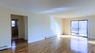 This single-floor space presents a bright, open living area defined by polished hardwood floors that run throughout, reflecting natural light and adding warmth to the interior. The walls are finished in a clean, neutral off-white tone, creating a versatile backdrop well-suited to a variety of furnishing styles. A large window on the far wall allows generous amounts of natural light to pour into the space, enhancing its airy and welcoming atmosphere. Baseboard heating units line the walls, providing efficient climate control while maintaining the clean aesthetic of the room. The layout is open and unobstructed, offering flexible arrangement possibilities. A doorway visible on the left side of the space leads to an adjoining area, suggesting additional rooms beyond the main living zone. The smooth, white ceiling complements the light-toned walls, contributing to the overall sense of openness. The space is well-maintained and move-in ready, with its combination of hardwood flooring, neutral finishes, and ample natural light making it a practical and comfortable living environment.