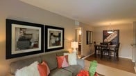 This thoughtfully designed single-floor residence offers a seamless and inviting living experience. The open-concept layout connects the living and dining areas, creating a sense of spaciousness and ease of flow throughout the home. The living area is anchored by a comfortable seating arrangement and features warm, neutral-toned walls that complement the hardwood-style flooring running through the space. Large, bold-framed wall art adds a sophisticated, contemporary touch to the room, while a classic table lamp provides warm ambient lighting. The dining area is conveniently positioned adjacent to the living space, featuring a dark-toned dining set that contrasts elegantly with the lighter surroundings. A hallway connects the main living areas to the rest of the home, hinting at additional rooms beyond. Recessed vents and clean ceiling lines reflect a practical yet polished interior finish. The overall ambiance is warm, modern, and welcoming, with a cohesive color palette of neutral beiges and grays accented by pops of coral and white in the soft furnishings. This space strikes a balance between comfort and style, making it an ideal environment for both relaxing and entertaining.