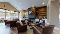 This well-appointed single-floor community space makes a strong first impression with its tiled entryway, featuring a geometric inlay pattern and tall, grid-paned French doors that frame the transition into the main lounge. The reception area includes a handsome reclaimed wood desk, setting a warm, rustic-modern tone carried throughout the space.

The central lounge is anchored by a dramatic floor-to-ceiling reclaimed wood accent wall, housing a sleek linear gas fireplace beneath a wall-mounted television. Rich brown leather armchairs are arranged around a round wooden coffee table, creating an inviting and comfortable gathering area. Soaring vaulted ceilings with recessed lighting enhance the sense of openness, while pendant lighting adds a layered warmth.

A separate kitchenette area features a peninsula counter with bar stools, stainless steel appliances including a full-size refrigerator, a microwave, and a sink — all complemented by soft blue-grey walls and pendant lighting. Adjacent to the lounge, a bright windowed area offers a row of high-top communal tables with industrial-style stools, bathed in abundant natural light from a sweeping bank of large windows overlooking the outdoors.

Throughout, warm hardwood flooring ties the various zones together, creating a cohesive and functional space well-suited for socializing, working, or relaxing.