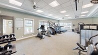Located at 3600 Corriere Rd in Easton, PA, this fitness center offers a thoughtfully designed exercise room that provides a comprehensive workout environment. The space features a drop-panel ceiling with recessed lighting and dual ceiling fans for optimal air circulation and comfort during workouts.

Natural light fills the room through multiple large windows, creating a bright and welcoming atmosphere. The soft sage-green walls, accented with white trim, contribute to a clean and motivating aesthetic. Durable, neutral-toned carpet tile covers the floor throughout.

The cardio zone is well-stocked with treadmills, elliptical machines, stair climbers, and stationary bikes — suitable for a wide range of aerobic workouts. The strength training area is equally impressive, featuring multiple SportsArt cable and weight machines designed to target various muscle groups. Full-length mirrors line one wall, enhancing the sense of space while allowing users to monitor their form.

A wall-mounted television is positioned for visibility across the room, adding an entertainment option during workouts. The overall layout is organized and spacious, allowing for comfortable movement between equipment. This fitness center delivers a well-rounded facility suitable for both cardio and strength training routines.