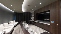 This elegantly designed private home theater delivers a premium cinematic experience through a seamless blend of upscale materials and thoughtful architectural details. The room is anchored by a large flat-screen display set within a floor-to-ceiling wall of rich, dark wood cabinetry, which provides generous media storage beneath a clean, continuous countertop surface. Tiered seating is arranged in a curved configuration, with plush white recliners offering unobstructed sightlines to the screen from every position. A wide, neutral-toned striped area rug grounds the seating area and adds a layer of texture to the refined interior. The ceiling is a standout architectural feature, showcasing a sweeping coved design with recessed lighting that casts a warm, layered glow throughout the space. Full-length dark drapery lines the walls, enhancing acoustics while contributing to the room's immersive, theater-like atmosphere. Subtle wall sconces provide gentle accent lighting, and a decorative floral arrangement adds a refined finishing touch. The overall ambiance is one of understated luxury — dark, dramatic, and purposefully designed for comfort and visual performance.