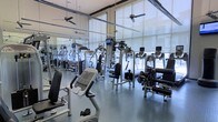 This single-floor fitness center offers a comprehensive workout environment designed to accommodate a wide range of exercise needs. The space is defined by its floor-to-ceiling windows, which line the perimeter and flood the interior with abundant natural light while providing views of the surrounding exterior. Sheer roller shades allow for light control without sacrificing the open, airy feel of the room.

The facility is outfitted with a robust selection of commercial-grade strength training machines and cardiovascular equipment, including cable machines, multi-functional weight stations, and cardio units such as stationary bikes and treadmills. Equipment is thoughtfully arranged to allow for ease of movement throughout the space. Full-length mirrored walls visually expand the room and provide functional feedback during workouts.

Overhead, multiple ceiling fans are suspended from the high ceiling, promoting air circulation throughout. The durable rubberized flooring provides both safety and practicality underfoot. Mounted monitors are integrated into select equipment, enhancing the user experience. Overall, the space conveys a clean, modern aesthetic suited for a high-quality fitness facility.