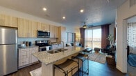 Located in Draper, Utah, this contemporary single-floor residence offers a thoughtfully designed open-concept layout that seamlessly connects the kitchen, dining, and living areas. The kitchen is outfitted with light wood cabinetry extending to the ceiling, stainless steel appliances including a refrigerator, electric range, and over-the-range microwave, as well as a granite-topped island with an integrated sink and bar seating for two — ideal for casual dining or entertaining. Rich, dark hardwood-style flooring runs throughout the main living areas, grounding the space with warmth and continuity. The living room is bathed in natural light from a large sliding glass door that opens to a private balcony, offering open views of the surrounding Draper landscape. A ceiling fan adds comfort to the space. The bedroom is generously sized, featuring large windows with plantation-style blinds that invite ample natural light, soft carpeting, and a ceiling fan. The bathroom continues the home's cohesive aesthetic with light wood cabinetry, a dual-sink granite vanity, a large mirror, and hardwood-style flooring. Recessed lighting and a neutral color palette throughout create a clean, modern ambiance that is both functional and inviting.