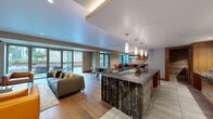 This thoughtfully designed single-floor amenity space offers an expansive, open-concept layout that seamlessly connects multiple functional areas. The entryway greets visitors with light-toned large-format tile flooring and textured wall paneling, setting a refined tone throughout. The gourmet kitchen is a standout centerpiece, featuring a dramatic black-and-white veined marble backsplash, rich walnut cabinetry, dark granite countertops, a substantial island with bar seating, pendant lighting, and stainless steel appliances. A secondary prep kitchen nearby provides additional functionality with matching dark stone counters and wood-plank tile walls. The open living area flows naturally from the kitchen, offering wide-plank hardwood floors, recessed and cove lighting, and floor-to-ceiling glass doors that connect the interior to the outdoor terrace. The lounge is anchored by a large wall-mounted display, wood media cabinetry, and a mix of contemporary seating. A dedicated dining area accommodates a generous table beneath a striking branching chandelier, enhanced by natural light from large windows. A game table area adds a social dimension to the space. Outside, a well-appointed courtyard features manicured planters with ornamental grasses, wood-clad bench seating, an outdoor kitchen, and open-air gathering areas framed by the surrounding urban architecture.