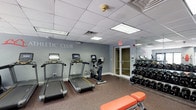 This athletic club is a dedicated fitness facility designed to provide residents or guests with a comprehensive workout space. The room features a drop ceiling with recessed fluorescent panel lighting that ensures the space is well-lit throughout. The walls are painted a cool grey-blue tone, with the words "LETIC CLUB" prominently displayed, indicating the space is part of a larger branded athletic club amenity.

The flooring consists of durable, dark rubber gym matting, ideal for high-traffic fitness use and equipment support. Along one wall, two treadmills and an upright stationary bike are neatly arranged, each equipped with individual digital display consoles for tracking workout metrics. A full-length mirror lines the opposite wall, visually expanding the space and allowing users to monitor their form during exercise.

A well-stocked dumbbell rack holds an organized selection of free weights, catering to strength training needs. An adjustable workout bench is positioned in the foreground. Access to the facility is provided through a set of glass double doors, allowing natural light to filter in. An emergency exit sign and fire detection equipment reflect the room's adherence to safety standards.
