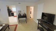 This single-floor residence offers a comfortable and functional layout with a cohesive, neutral color palette throughout. The main living area is carpeted and open, providing a versatile space suitable for both relaxing and working. A small writing desk with a table lamp and decorative accents occupies one corner, adding a practical workspace element to the room. A media console with a flat-screen television is positioned along the opposite wall, making the living space well-suited for everyday use. The room flows naturally toward a hallway that leads to additional areas of the home, with a glimpse of a bathroom featuring a towel rack and warm lighting visible in the distance. To one side, the kitchen is partially visible and features white cabinetry, dark appliances, and a window that allows natural light to filter into the space. A patterned area rug anchors the living area, adding a touch of warmth and visual interest to the light-toned carpet. The space is finished with clean white walls, white trim, and standard ceiling vents, conveying a tidy and well-maintained interior. Overall, the layout is efficient and straightforward, offering a welcoming single-level living environment.
