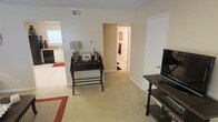 This single-floor residence offers a comfortable and functional layout with a cohesive, neutral color palette throughout. The main living area is carpeted and open, providing a versatile space suitable for both relaxing and working. A small writing desk with a table lamp and decorative accents occupies one corner, adding a practical workspace element to the room. A media console with a flat-screen television is positioned along the opposite wall, making the living space well-suited for everyday use. The room flows naturally toward a hallway that leads to additional areas of the home, with a glimpse of a bathroom featuring a towel rack and warm lighting visible in the distance. To one side, the kitchen is partially visible and features white cabinetry, dark appliances, and a window that allows natural light to filter into the space. A patterned area rug anchors the living area, adding a touch of warmth and visual interest to the light-toned carpet. The space is finished with clean white walls, white trim, and standard ceiling vents, conveying a tidy and well-maintained interior. Overall, the layout is efficient and straightforward, offering a welcoming single-level living environment.
