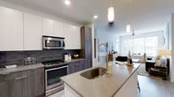 This single-floor space showcases a contemporary open-concept design, anchored by a well-appointed kitchen that serves as the heart of the home. The kitchen features a large island with a light quartz countertop and an undermount sink equipped with a high-arc spring-style faucet, offering both functionality and style. Two pendant lights hang above, providing focused task lighting that complements the recessed ceiling lights throughout.

Cabinetry combines crisp white upper cabinets with wood-toned lower cabinets, creating a two-tone aesthetic that feels both modern and warm. A dark subway tile backsplash adds texture and contrast behind the stainless steel range and over-the-range microwave, reinforcing the space's contemporary character. A tall pantry-style cabinet provides generous storage.

The kitchen flows naturally into the living area, which benefits from abundant natural light streaming through large windows. A ceiling fan is visible in the background, contributing to comfort and airflow. The overall ambiance is clean, bright, and cohesive, with a neutral palette and modern finishes throughout that give the space a polished, move-in-ready feel.