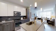 This single-floor space showcases a contemporary open-concept design, anchored by a well-appointed kitchen that serves as the heart of the home. The kitchen features a large island with a light quartz countertop and an undermount sink equipped with a high-arc spring-style faucet, offering both functionality and style. Two pendant lights hang above, providing focused task lighting that complements the recessed ceiling lights throughout.

Cabinetry combines crisp white upper cabinets with wood-toned lower cabinets, creating a two-tone aesthetic that feels both modern and warm. A dark subway tile backsplash adds texture and contrast behind the stainless steel range and over-the-range microwave, reinforcing the space's contemporary character. A tall pantry-style cabinet provides generous storage.

The kitchen flows naturally into the living area, which benefits from abundant natural light streaming through large windows. A ceiling fan is visible in the background, contributing to comfort and airflow. The overall ambiance is clean, bright, and cohesive, with a neutral palette and modern finishes throughout that give the space a polished, move-in-ready feel.